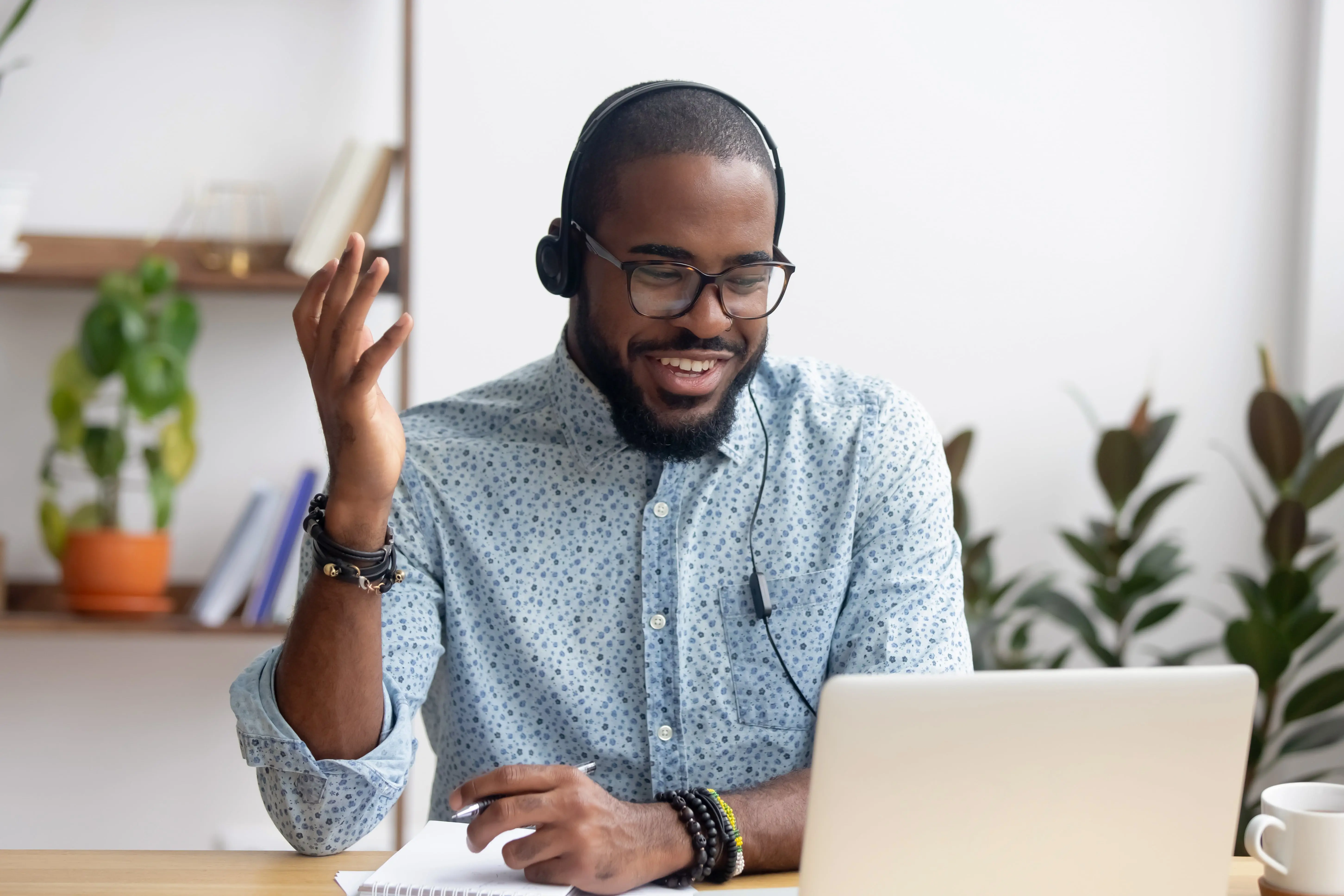 A man sat at a desk in a modern office wearing a blue shirt and glasses with a headset on talking to his laptop.