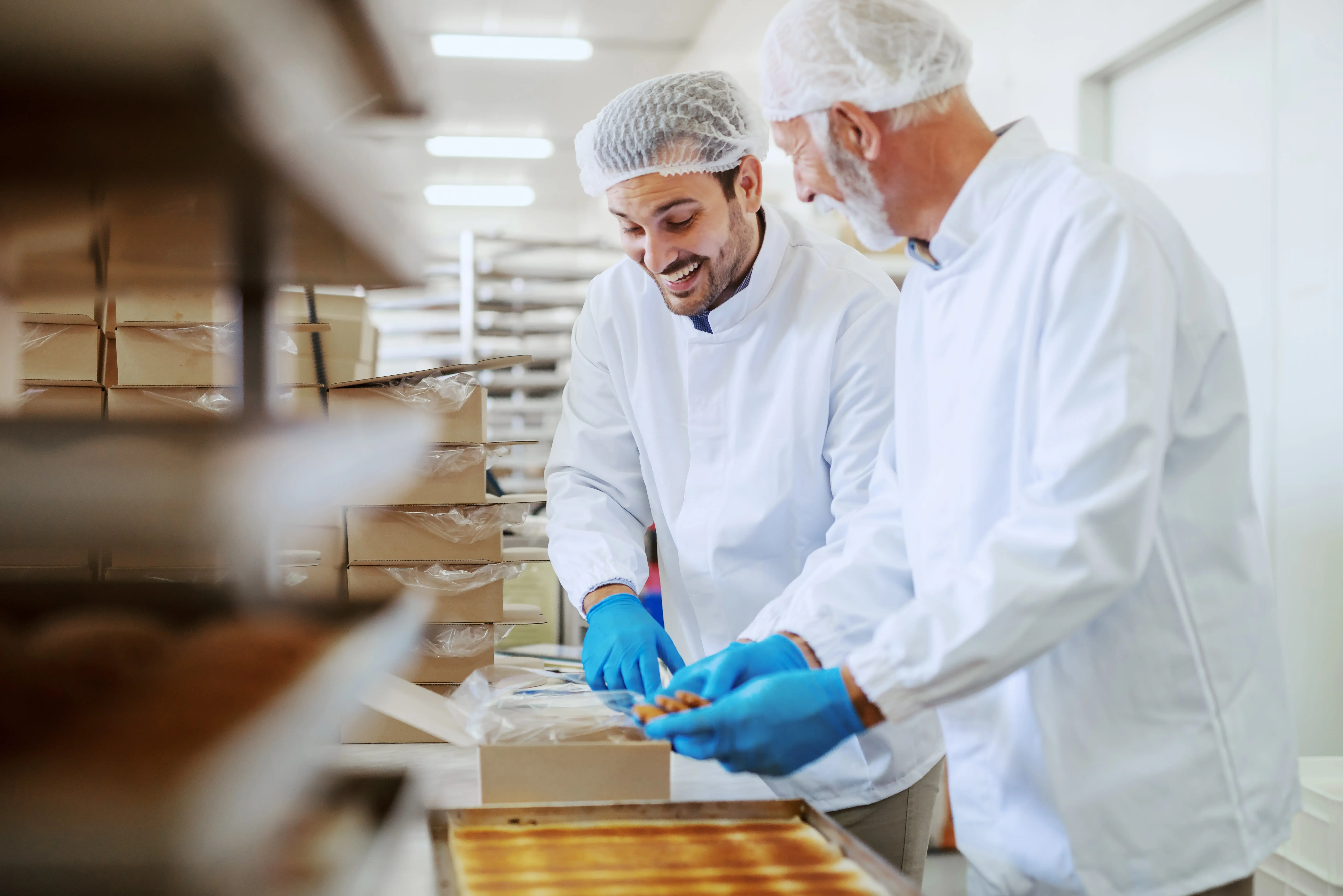Two workers in white lab coats, hairnets, and blue gloves are packaging baked goods in a commercial kitchen. They are smiling and working together while surrounded by stacks of boxes and trays.