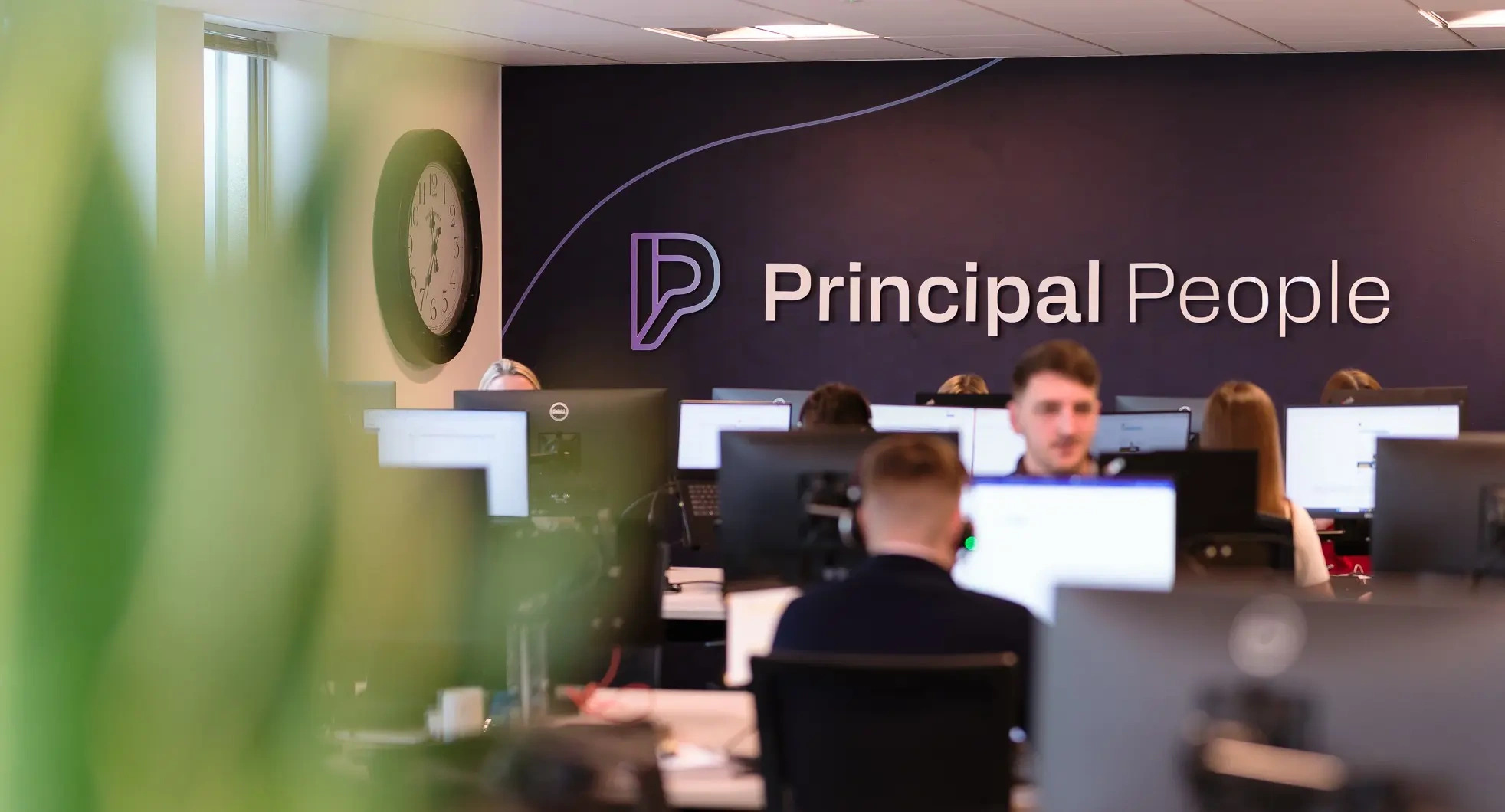 A group of professional people in a modern office with a large logo wall in the distance of the image.
