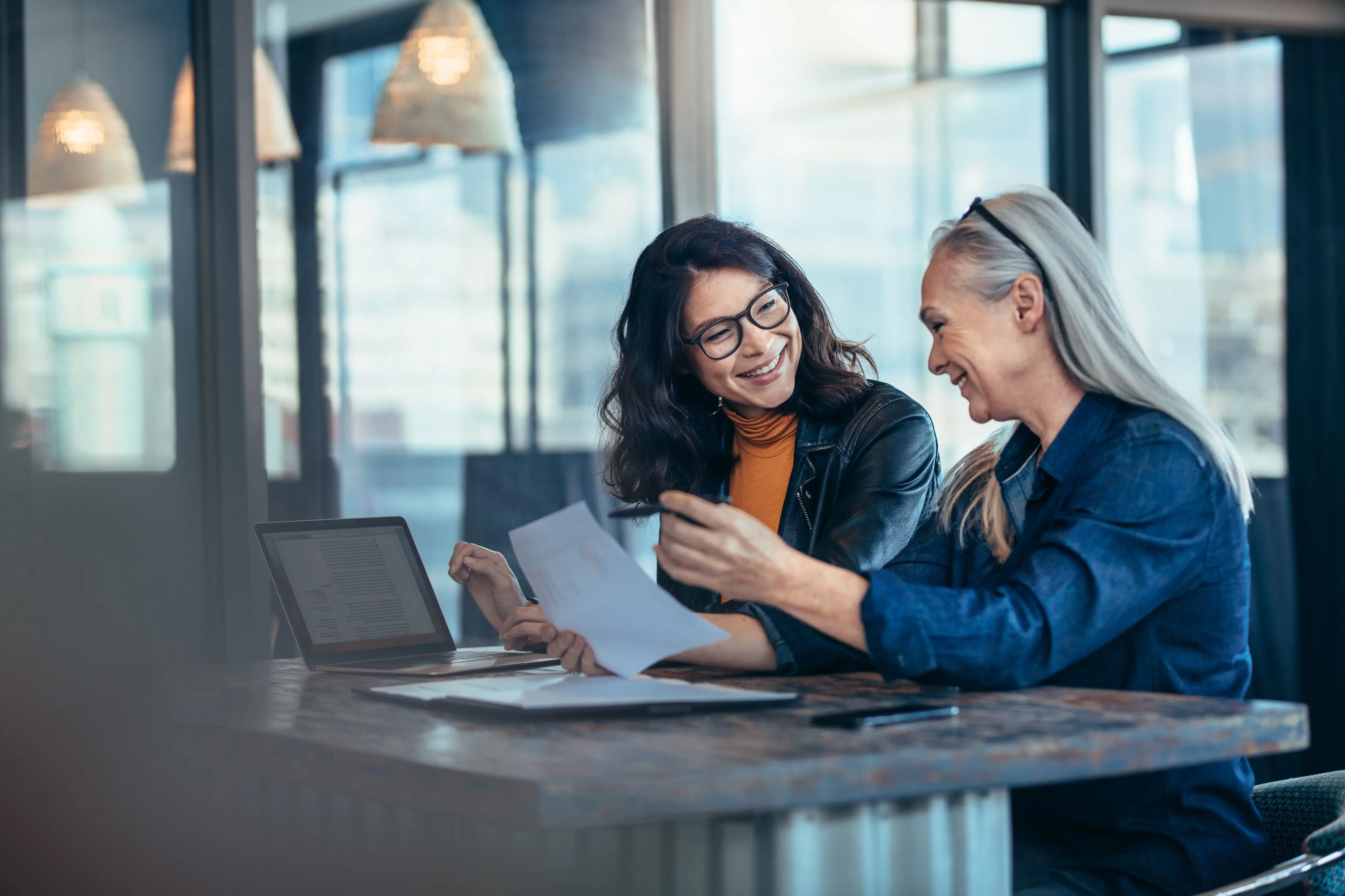 Two women speaking at a desk holding paper and one writing on a laptop