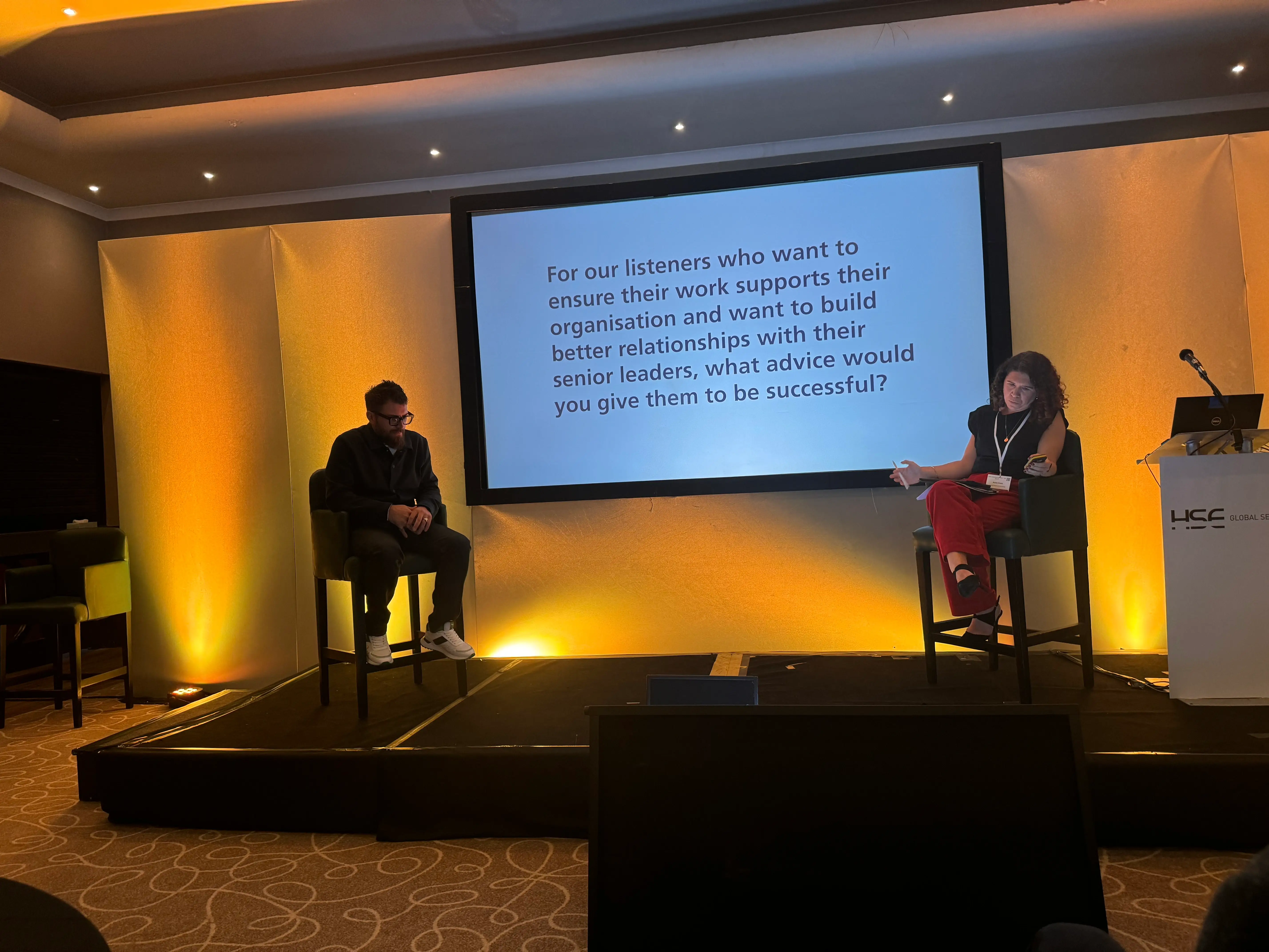 A male and a female in a dimly yellow light lit stage either side of a slideshow.