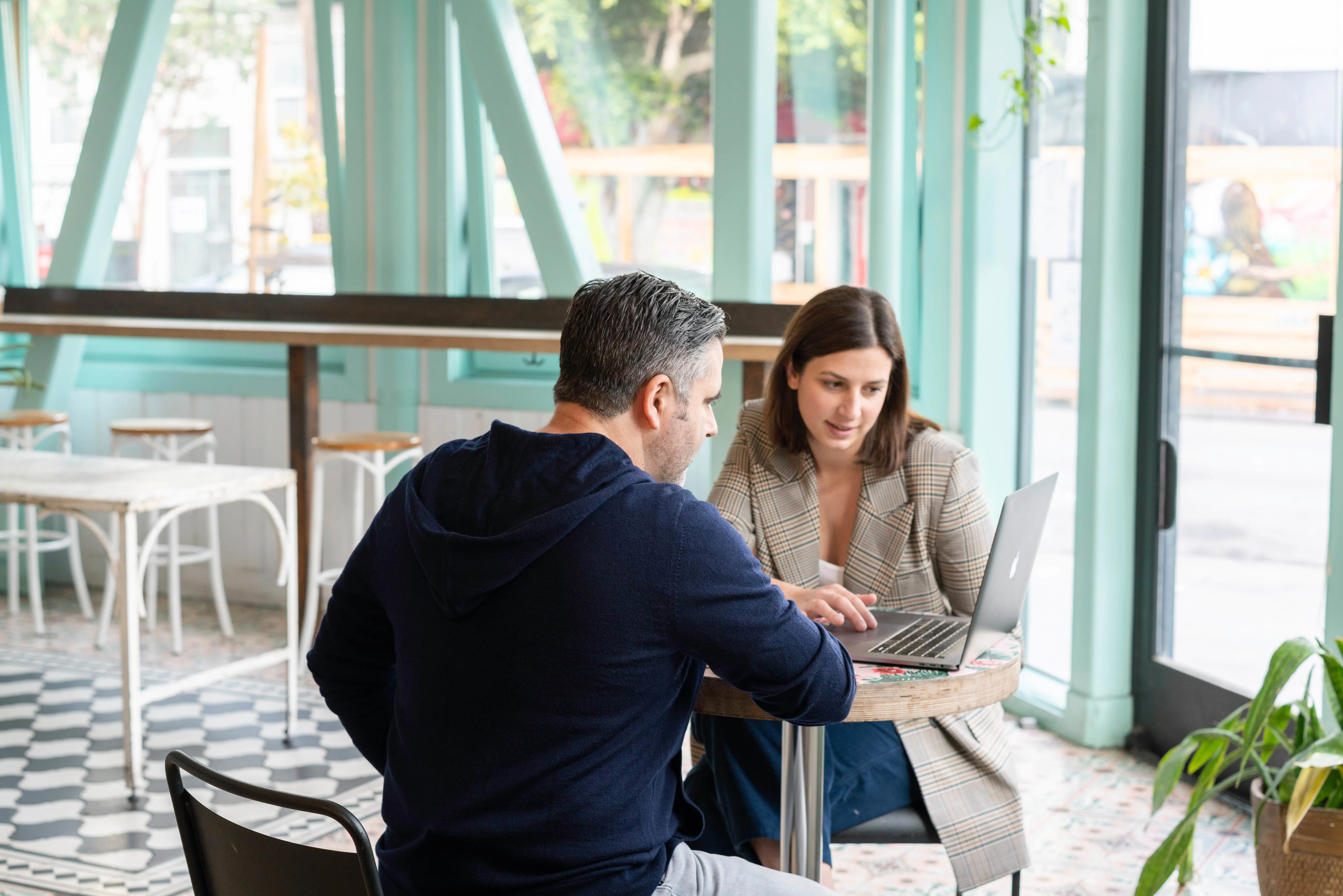 A man and woman sat at a round table in a cafe looking at a laptop.
