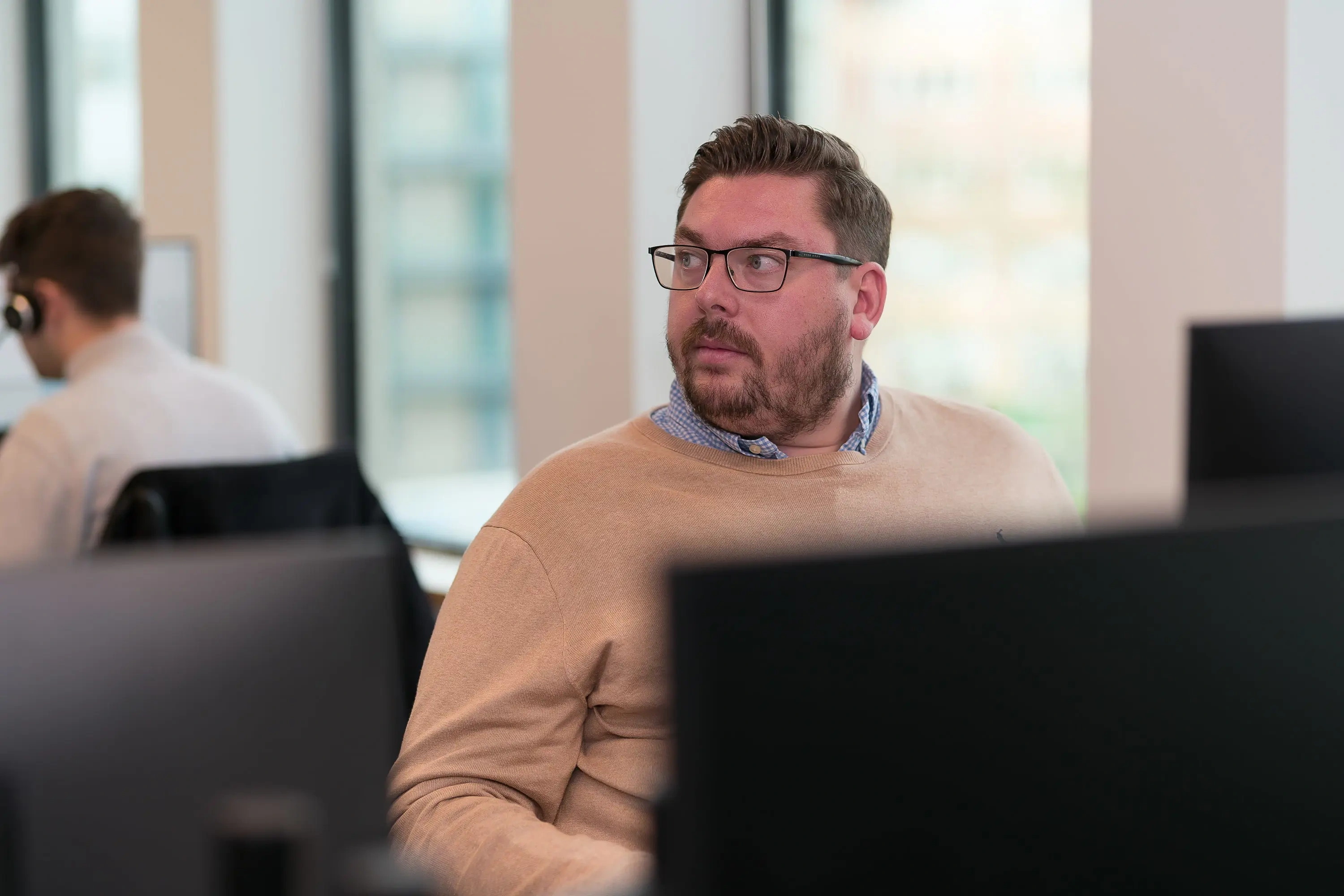 A man looking to the side sat at a desk in business casual clothes.
