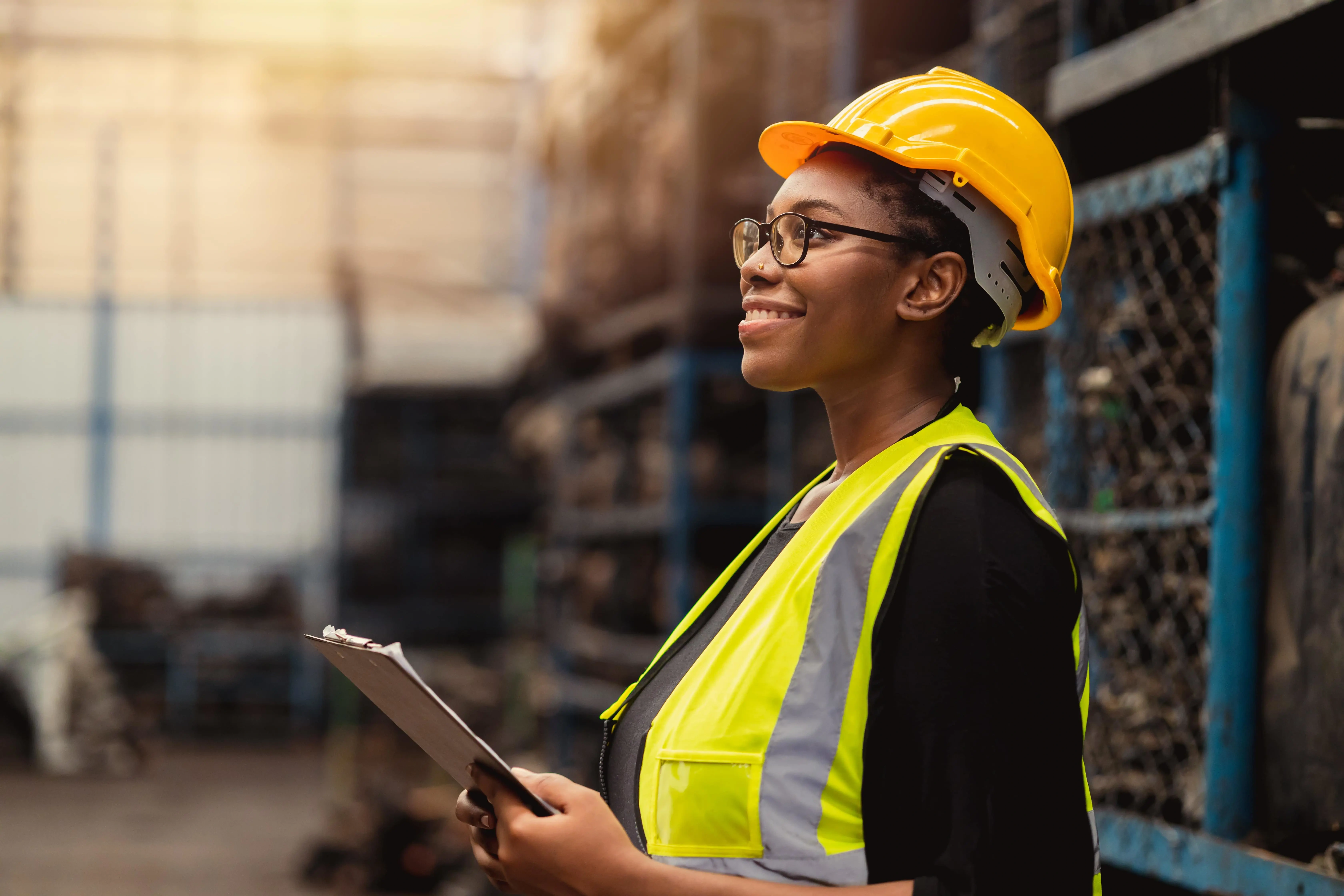 A woman wearing a yellow hard hat and safety vest holding a clipboard with her body facing the left.