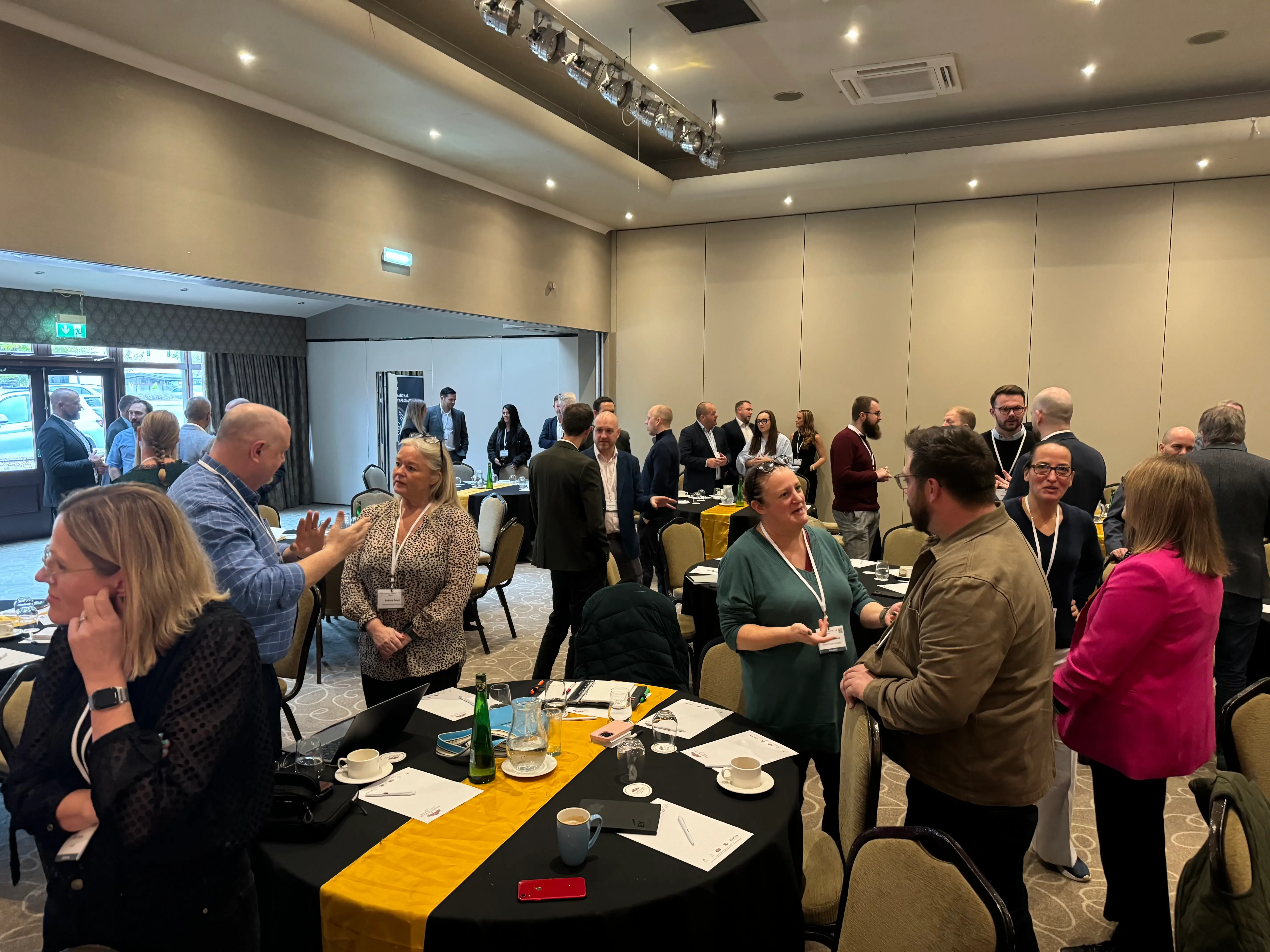 A large amount of people in a business event room stood around tables talking.