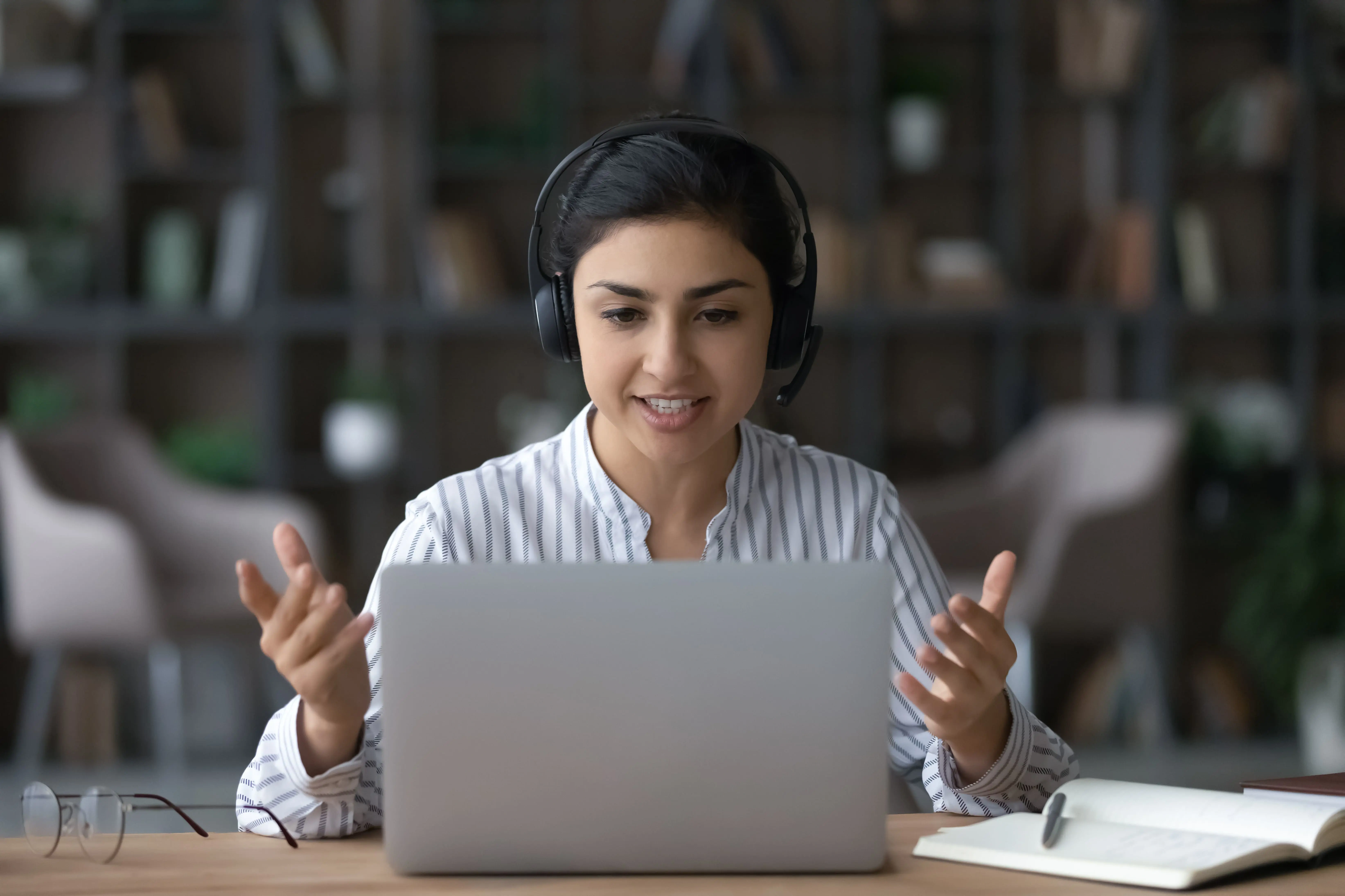 A young woman sat at a desk in a modern office space reading from a laptop wearing a headset and writing on a notepad.
